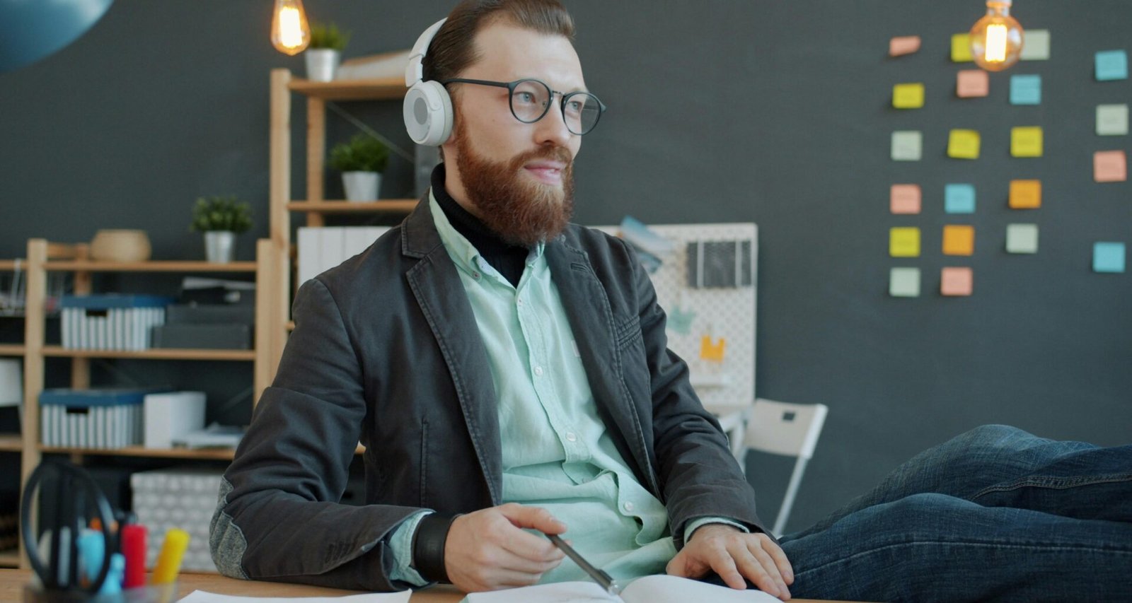 Mann mit Kopfhörern sitzt an einem Schreibtisch und arbeitet mit einem Notizbuch in einem modernen Büro