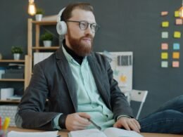 Mann mit Kopfhörern sitzt an einem Schreibtisch und arbeitet mit einem Notizbuch in einem modernen Büro