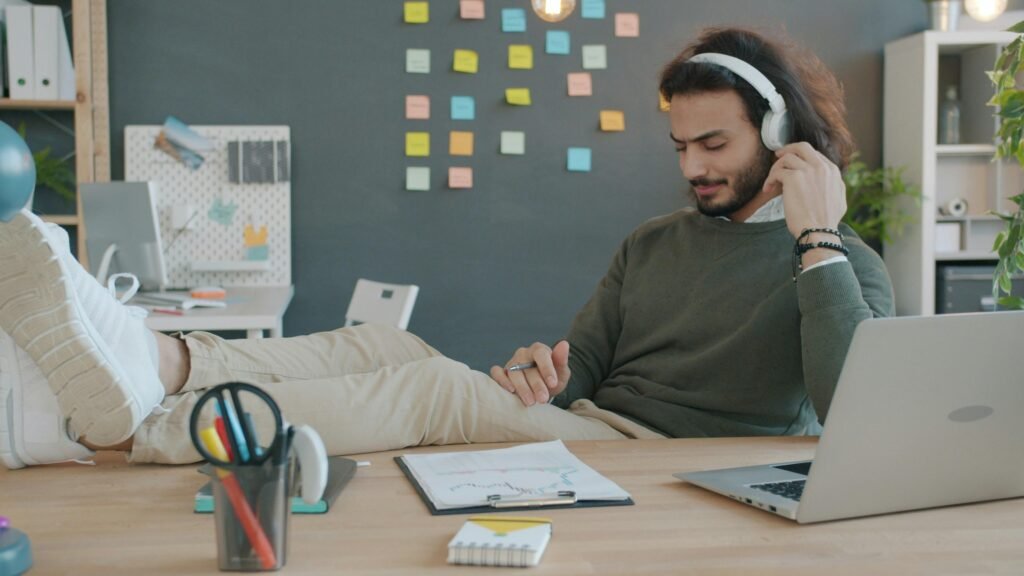 Mann trägt Kopfhörer und hört Musik am Schreibtisch während der Arbeit, moderner Arbeitsplatz mit Laptop und Notizbuch