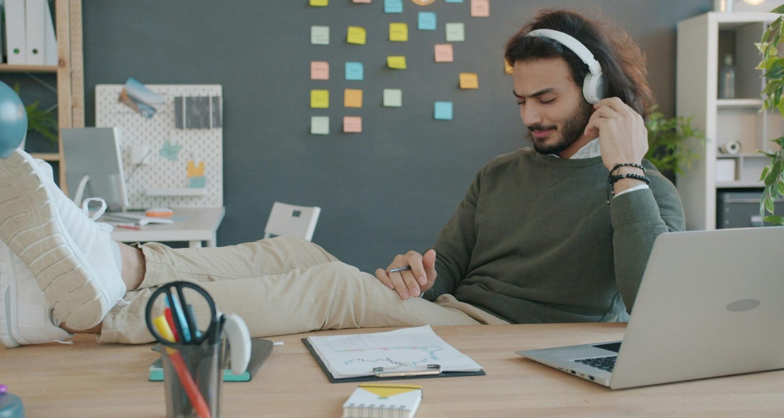 Mann trägt Kopfhörer und hört Musik am Schreibtisch während der Arbeit, moderner Arbeitsplatz mit Laptop und Notizbuch