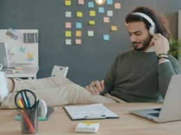 Mann sitzt am Schreibtisch und hört mit Kopfhörern Musik während der Arbeit, Fokus auf Technik und modernes Arbeiten