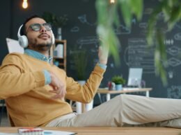Mann mit Kopfhörern spielt Luftgitarre an einem Schreibtisch in einem modernen Büro, Konzentration auf Musik