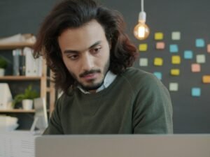 Mann mit langen Haaren arbeitet an einem Laptop in einem modernen Büro mit heller Einrichtung und großen Fenstern
