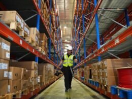 Mann in reflektierender Weste geht einen langen Lagergang entlang, umgeben von hohen Regalen mit Waren