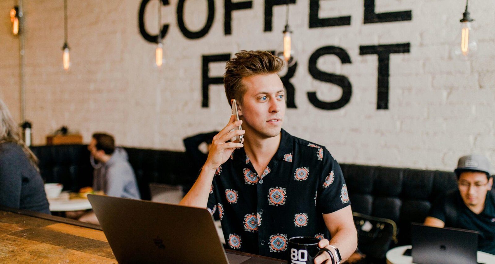 Mann hält ein Smartphone in der Hand und arbeitet gleichzeitig an einem MacBook an einem Schreibtisch