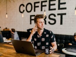 Mann hält ein Smartphone in der Hand und arbeitet gleichzeitig an einem MacBook an einem Schreibtisch