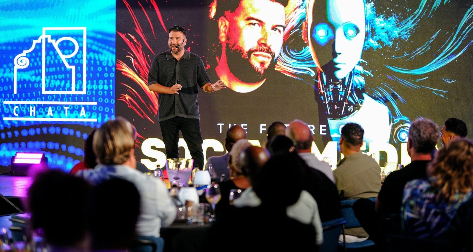 Ein Mann steht auf einer Bühne und hält eine Präsentation, hinter ihm eine große projizierte Roboter-Grafik als visueller Hintergrund.