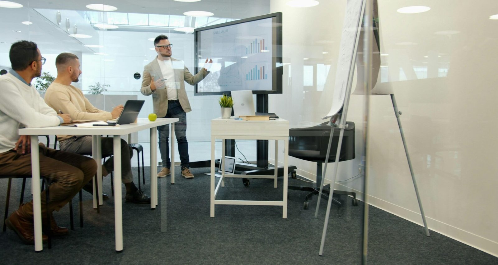 Ein Mann hält eine Präsentation vor seinen Kollegen in einem hellen, modernen Büro mit großen Fenstern und Monitoren.