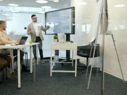 Ein Mann hält eine Präsentation vor seinen Kollegen in einem hellen, modernen Büro mit großen Fenstern und Monitoren.