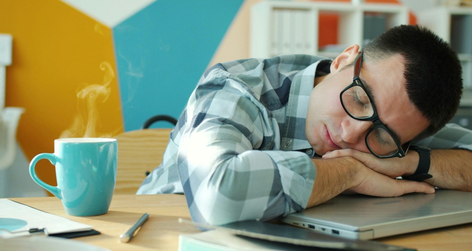 Ein Mann schläft an seinem Schreibtisch, neben ihm steht eine Kaffeetasse; Alltag im modernen Büro.