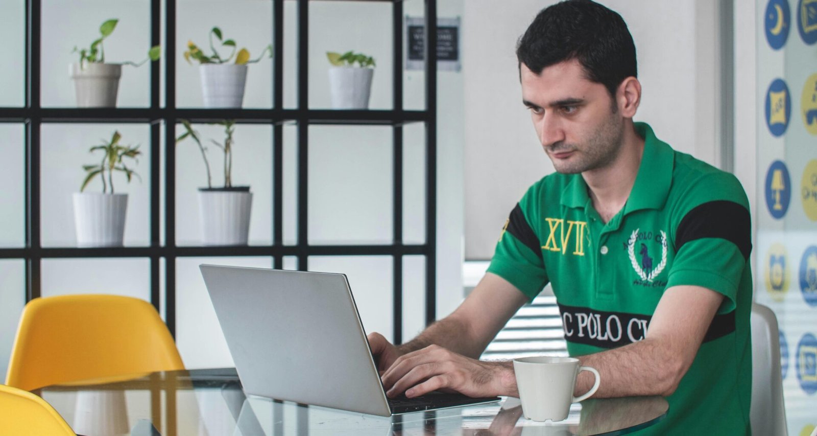 Mann sitzt auf einem grauen Metallstuhl und verwendet einen grauen Laptop an einem Schreibtisch in einem modernen Raum.