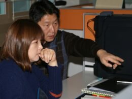 Ein Mann sitzt neben einer Frau, beide lehnen sich an einen weißen Tisch in einem hellen, modernen Büroraum.