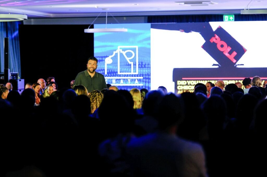 Mann spricht auf einer Bühne vor einem sitzenden Publikum, Lichtsituation in einem großen Saal, aufgenommen von Carlos Gil