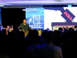 Mann spricht auf einer Bühne vor einem sitzenden Publikum, Lichtsituation in einem großen Saal, aufgenommen von Carlos Gil