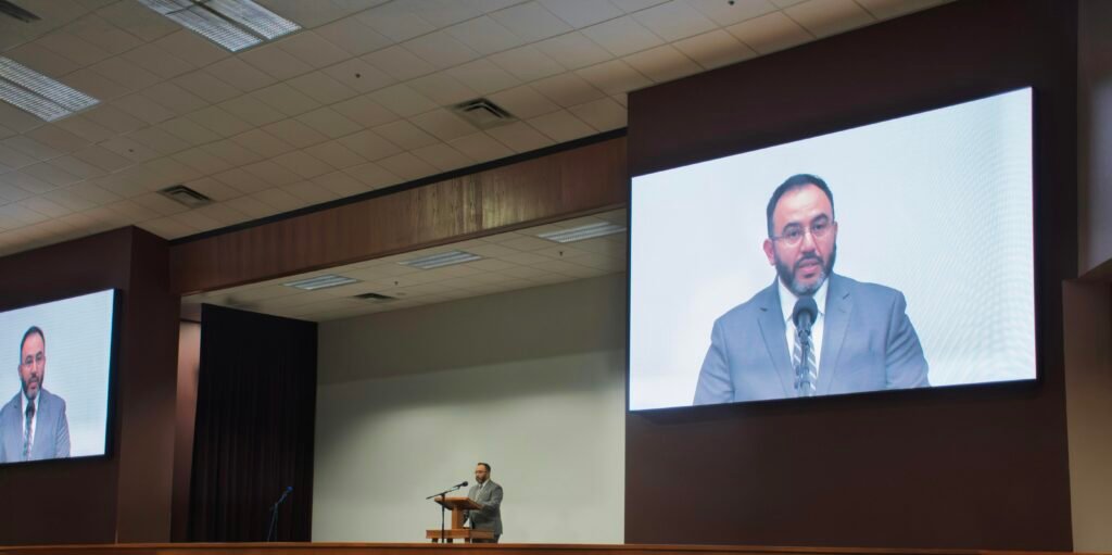 Ein Mann spricht an einem Podium, während sein Vortrag als Video auf einer großen Leinwand im Hintergrund angezeigt wird.