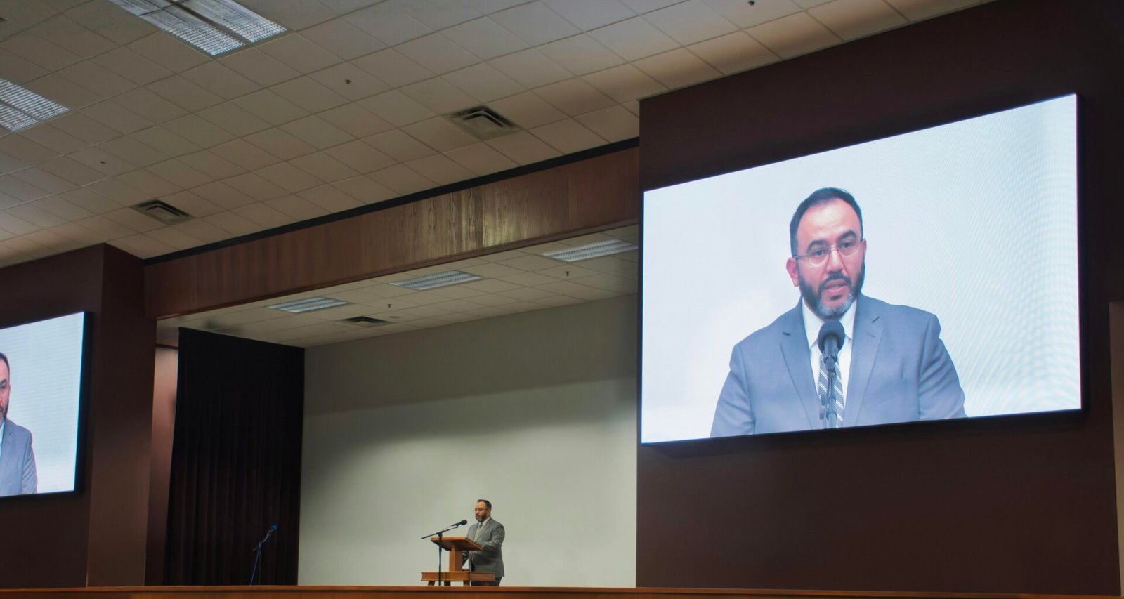 Ein Mann spricht an einem Podium, während sein Vortrag als Video auf einer großen Leinwand im Hintergrund angezeigt wird.
