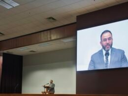 Ein Mann spricht an einem Podium, während sein Vortrag als Video auf einer großen Leinwand im Hintergrund angezeigt wird.
