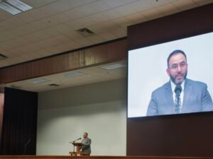 Ein Mann spricht an einem Podium, während sein Vortrag als Video auf einer großen Leinwand im Hintergrund angezeigt wird.