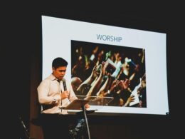 Ein Mann steht auf einer Bühne hinter einem Podium und präsentiert vor einem Publikum in einem Veranstaltungsraum.