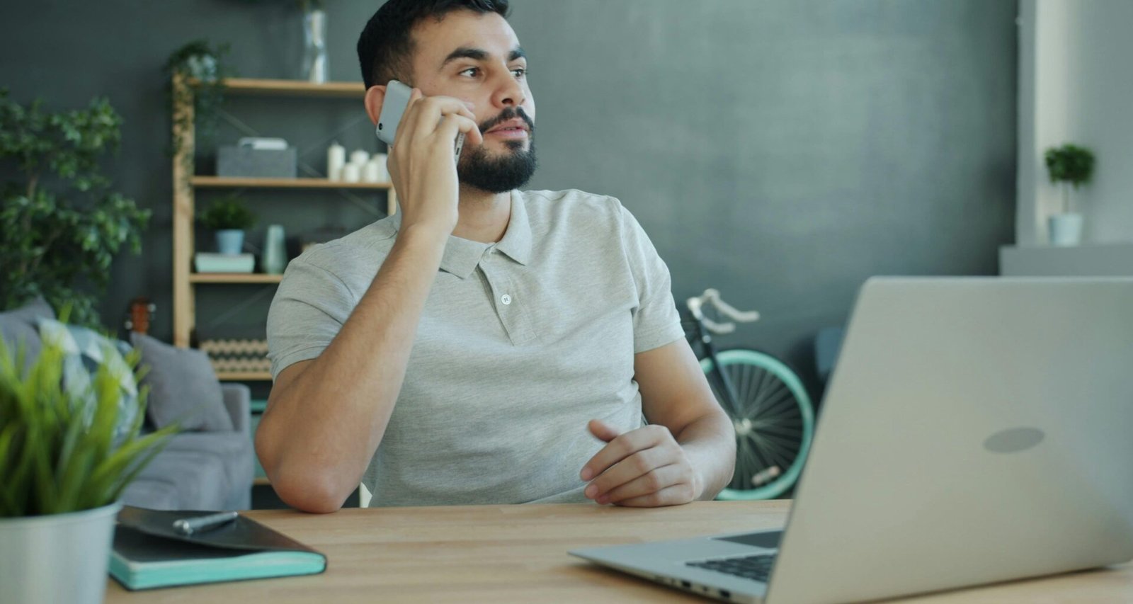 Mann telefoniert am Schreibtisch mit offenem Laptop in moderner Arbeitsumgebung bei Tageslicht.