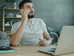 Mann telefoniert am Schreibtisch mit offenem Laptop in moderner Arbeitsumgebung bei Tageslicht.