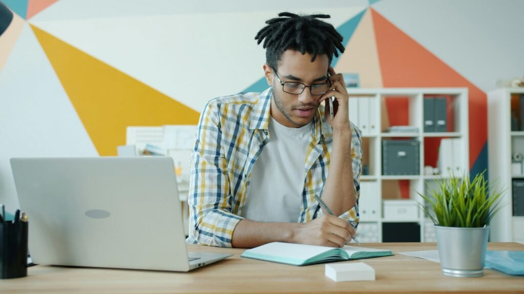 Mann telefoniert am Schreibtisch, arbeitet am Laptop und schreibt Notizen in ein Notizbuch im Büro
