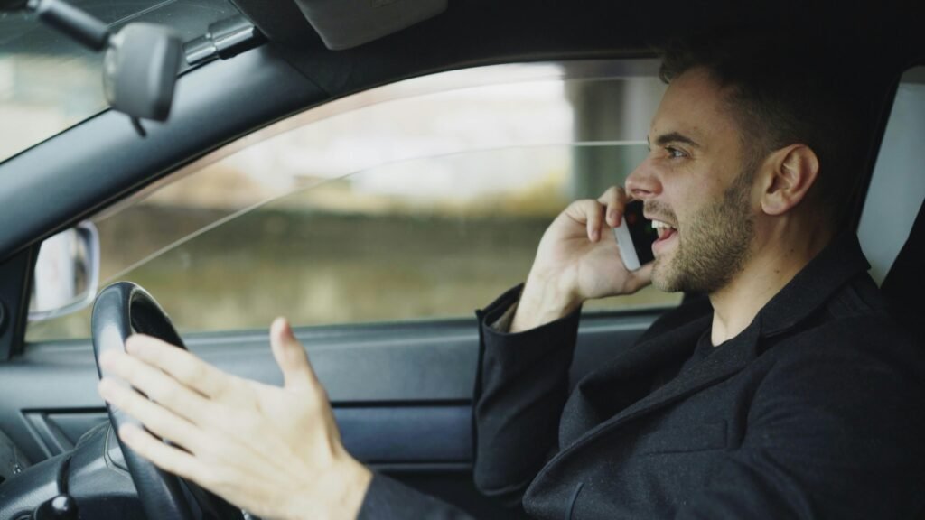 Ein Mann führt ein Telefonat mit dem Handy, während er ein Auto fährt, die Hände am Lenkrad und das Telefon am Ohr.