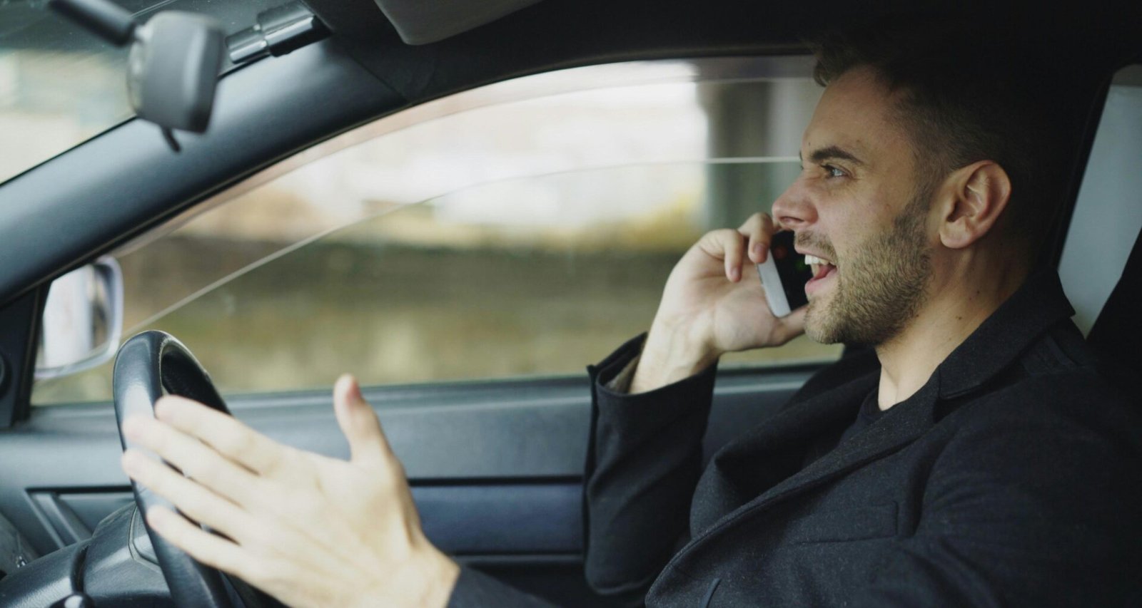 Ein Mann führt ein Telefonat mit dem Handy, während er ein Auto fährt, die Hände am Lenkrad und das Telefon am Ohr.