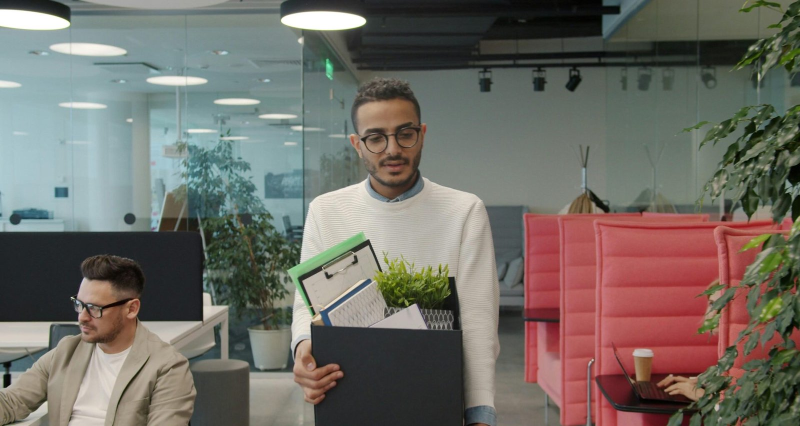 Mann trägt eine Kiste mit persönlichen Gegenständen durch ein modernes, helles Büro mit Glaswänden