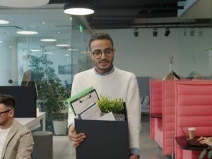 Mann trägt eine Kiste mit persönlichen Gegenständen durch ein modernes, helles Büro mit Glaswänden