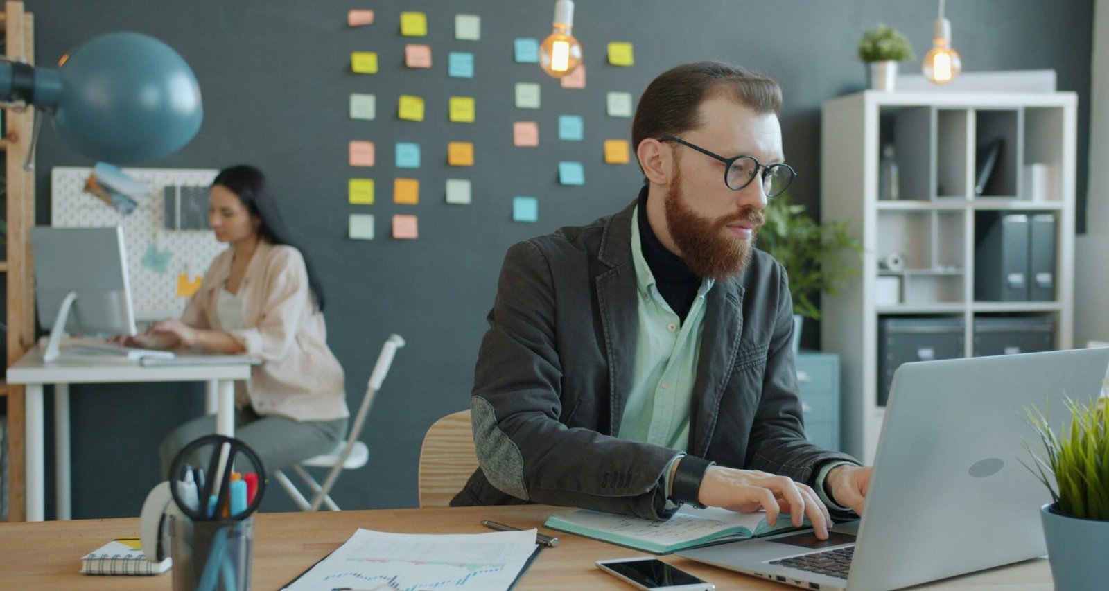 Ein Mann und eine Frau sitzen an Schreibtischen in einem modernen Büro mit Laptops und arbeiten zusammen an einem Projekt.