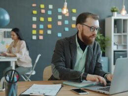 Ein Mann und eine Frau sitzen an Schreibtischen in einem modernen Büro mit Laptops und arbeiten zusammen an einem Projekt.