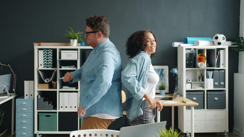 Ein Mann und eine Frau sitzen Rücken an Rücken an Schreibtischen im modernen Büro, mit Technik und Arbeitsmaterialien.
