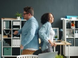 Ein Mann und eine Frau sitzen Rücken an Rücken an Schreibtischen im modernen Büro, mit Technik und Arbeitsmaterialien.