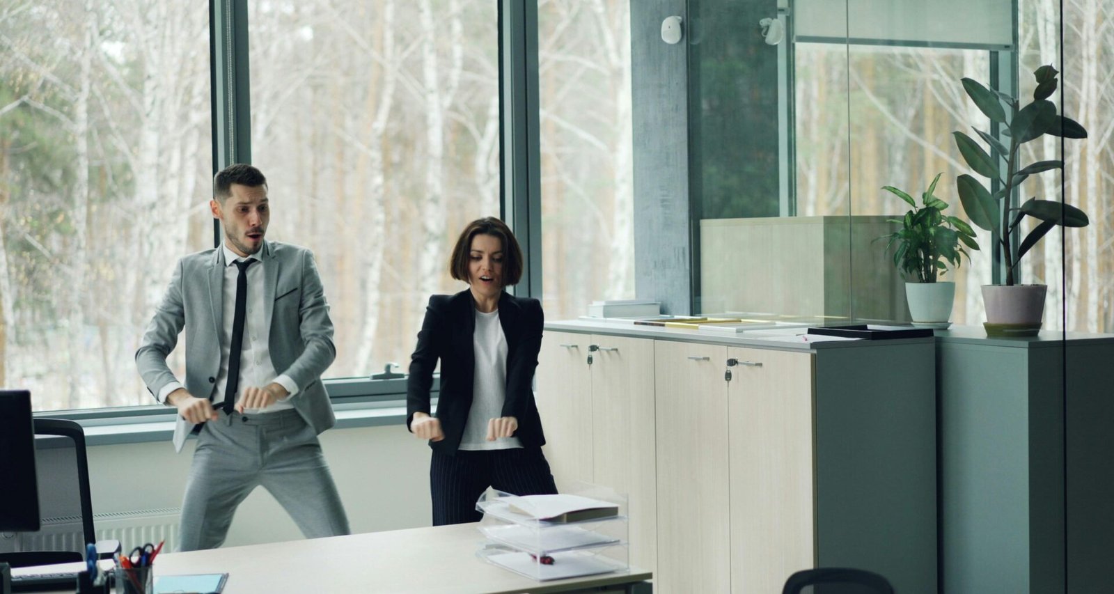 Ein Mann und eine Frau tanzen zusammen in einem hellen, modernen Büro mit Tischen und Computern im Hintergrund.