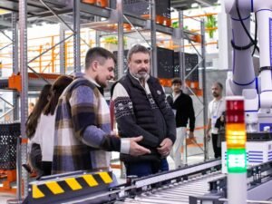 Mitarbeiter beobachten ein automatisiertes Förderbandsystem in einem modernen Lagerhaus mit zahlreichen Paketen und Regalen.