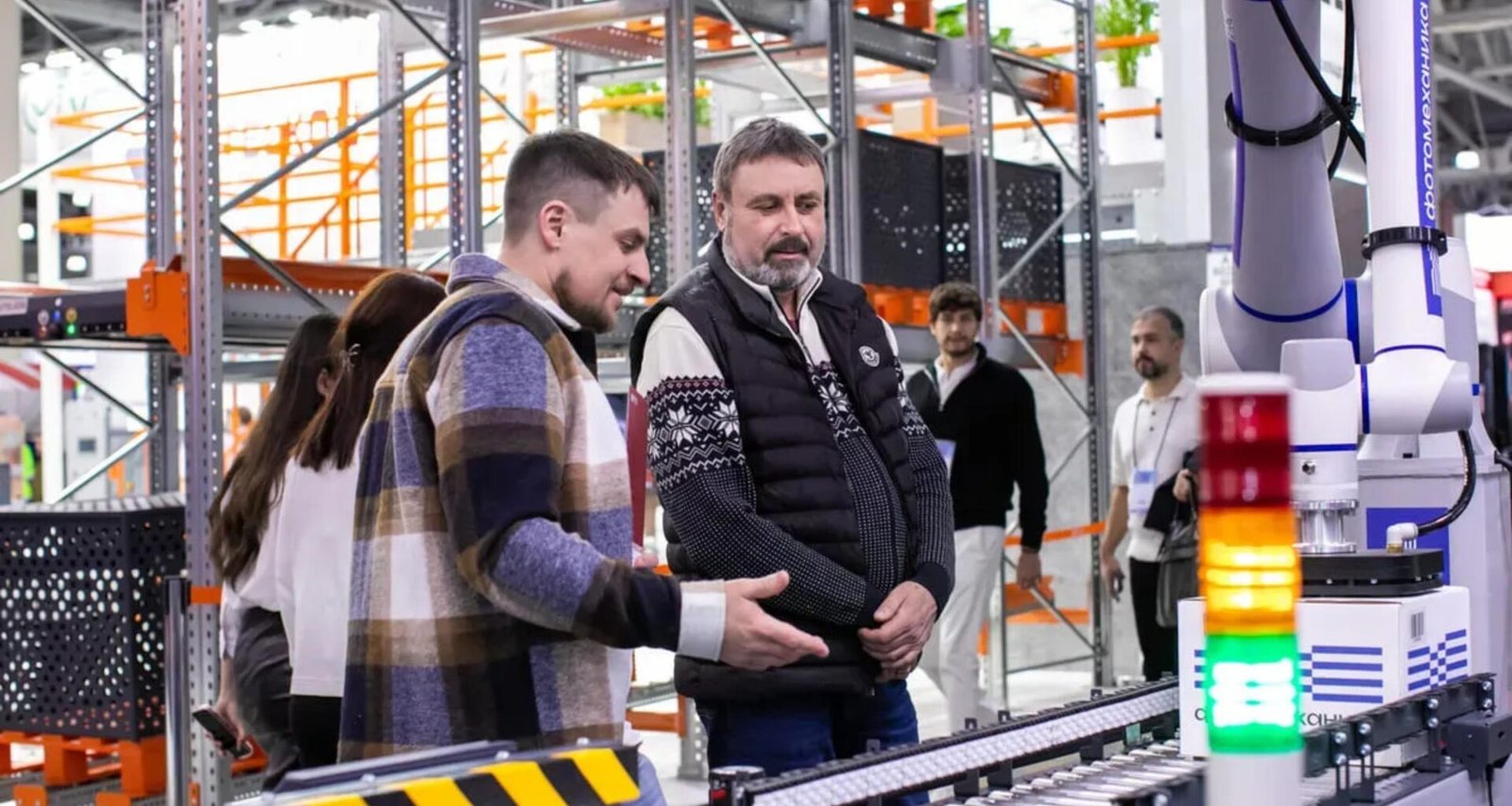 Mitarbeiter beobachten ein automatisiertes Förderbandsystem in einem großen Lagerhaus mit Regalen und Paketen.