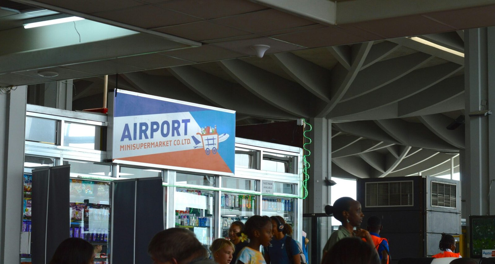 Personen stehen und gehen in einem hellen Flughafen-Terminal nahe einem großen Anzeigeschild für Abflüge und Gates.