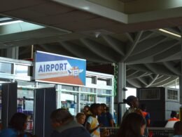 Personen stehen und gehen in einem hellen Flughafen-Terminal nahe einem großen Anzeigeschild für Abflüge und Gates.