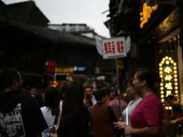 Menschen schlendern bei Dämmerung über eine gut besuchte Straße in der Innenstadt mit leuchtenden Schaufenstern.