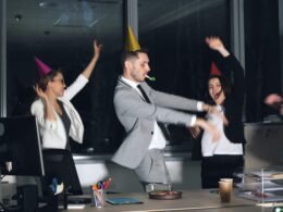 Mehrere Menschen tragen Partyhüte und tanzen in einem modernen Büro bei einer betrieblichen Feier.