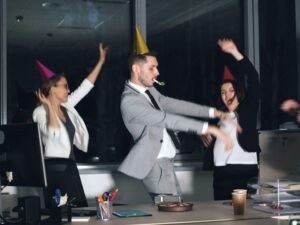 Mehrere Menschen tragen Partyhüte und tanzen in einem modernen Büro bei einer betrieblichen Feier.
