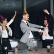 Mitarbeiter mit Partyhüten tanzen bei einer Bürofeier und feiern Erfolge am Arbeitsplatz