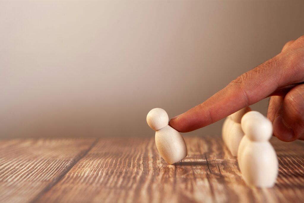 Ein Finger schiebt eine kleine Holzfigur vor einer Gruppe weiterer Figuren aus Holz auf einer hellen Oberfläche.