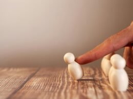 Ein Finger schiebt eine kleine Holzfigur vor einer Gruppe weiterer Figuren aus Holz auf einer hellen Oberfläche.