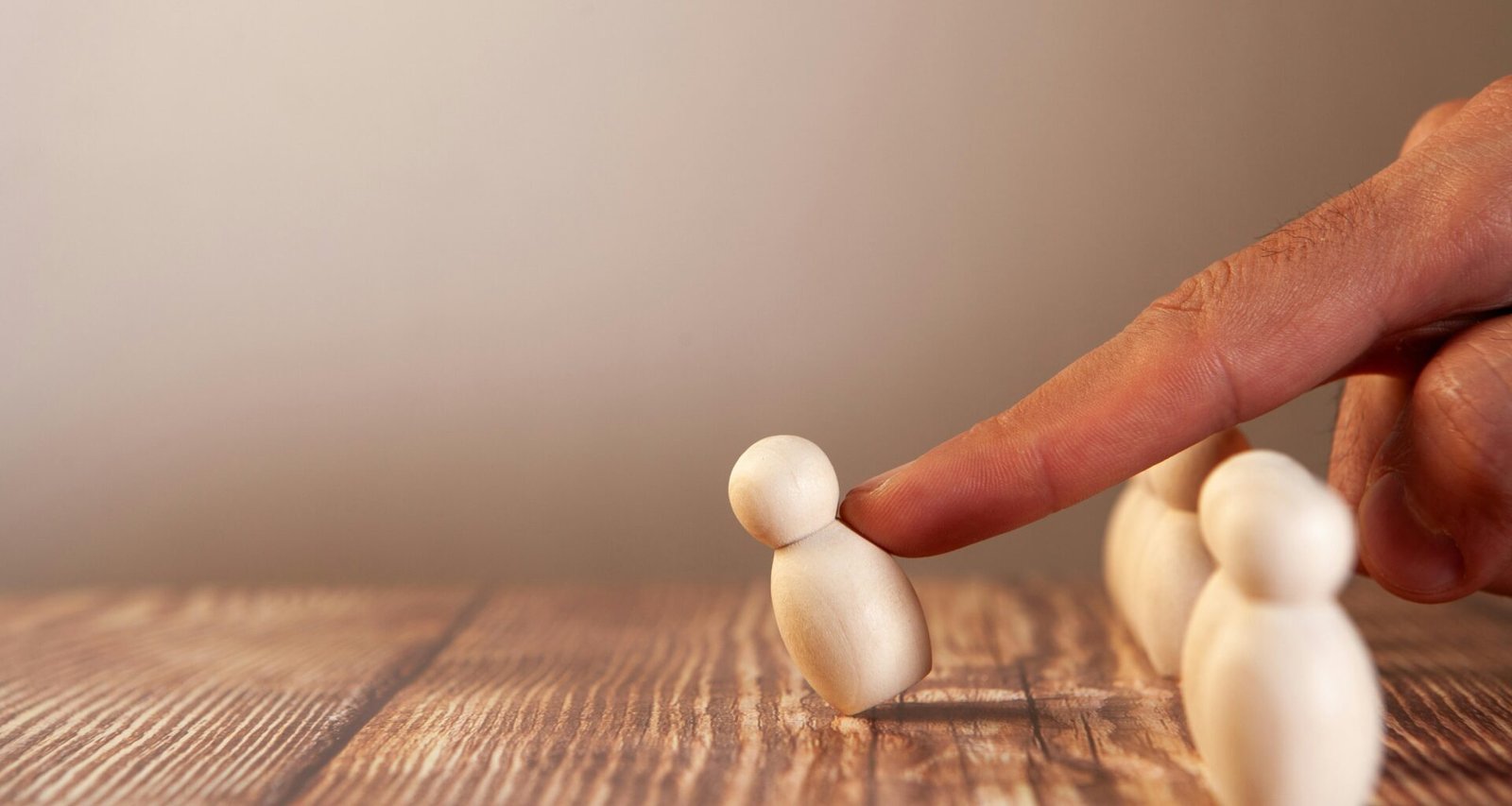 Eine Hand drückt eine kleine Holzfigur von einer Gruppe ähnlicher Figuren weg, Symbol für Gruppendynamik oder Entscheidungsprozesse.
