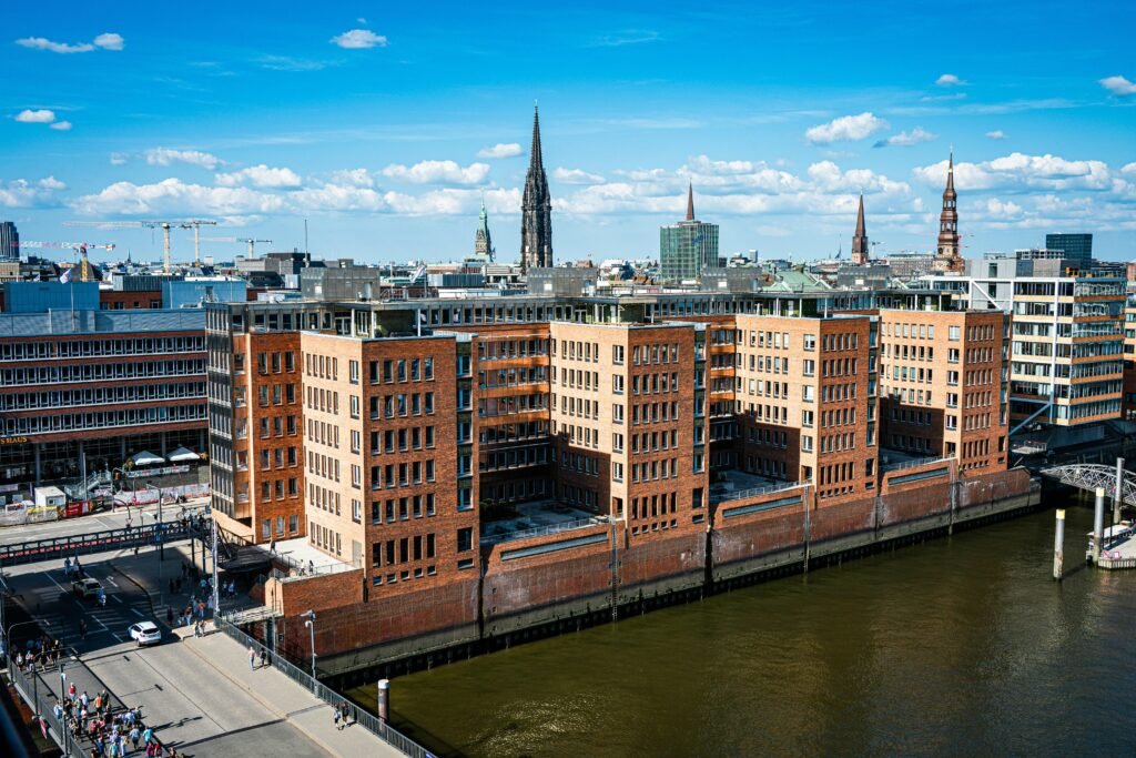Moderne Backsteinbauten stehen am Wasser, im Hintergrund erhebt sich die Skyline einer Stadt mit mehreren Hochhäusern.