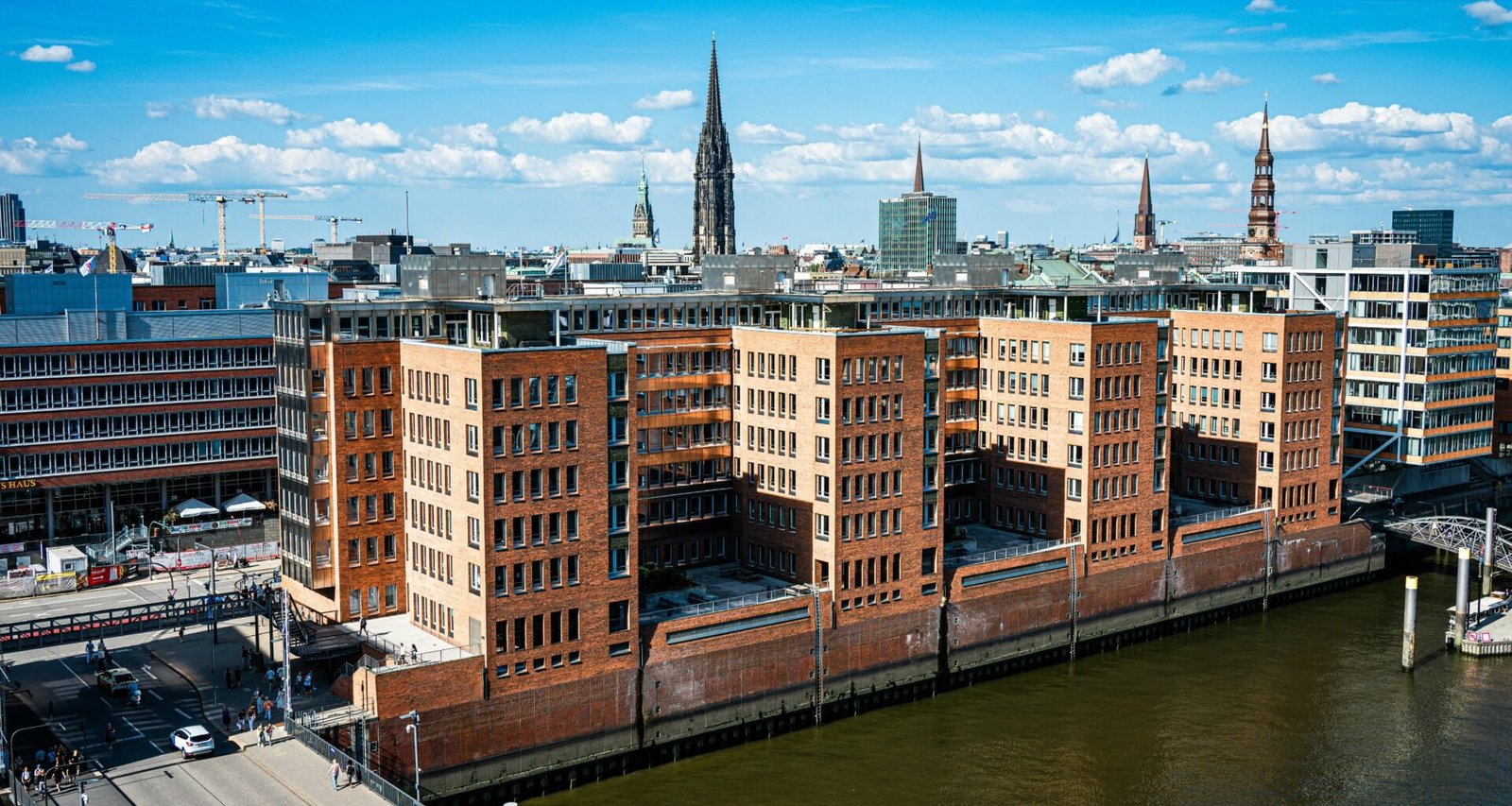 Moderne Backsteinbauten stehen am Wasser, im Hintergrund erhebt sich die Skyline einer Stadt mit mehreren Hochhäusern.