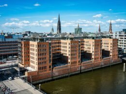 Moderne Backsteinbauten stehen am Wasser, im Hintergrund erhebt sich die Skyline einer Stadt mit mehreren Hochhäusern.