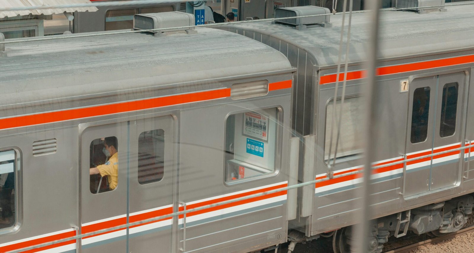 Moderner silberner Zug mit orangefarbener Linie steht am Bahnsteig einer Bahnhofsstation, aufgenommen von Reyhan Aviseno.